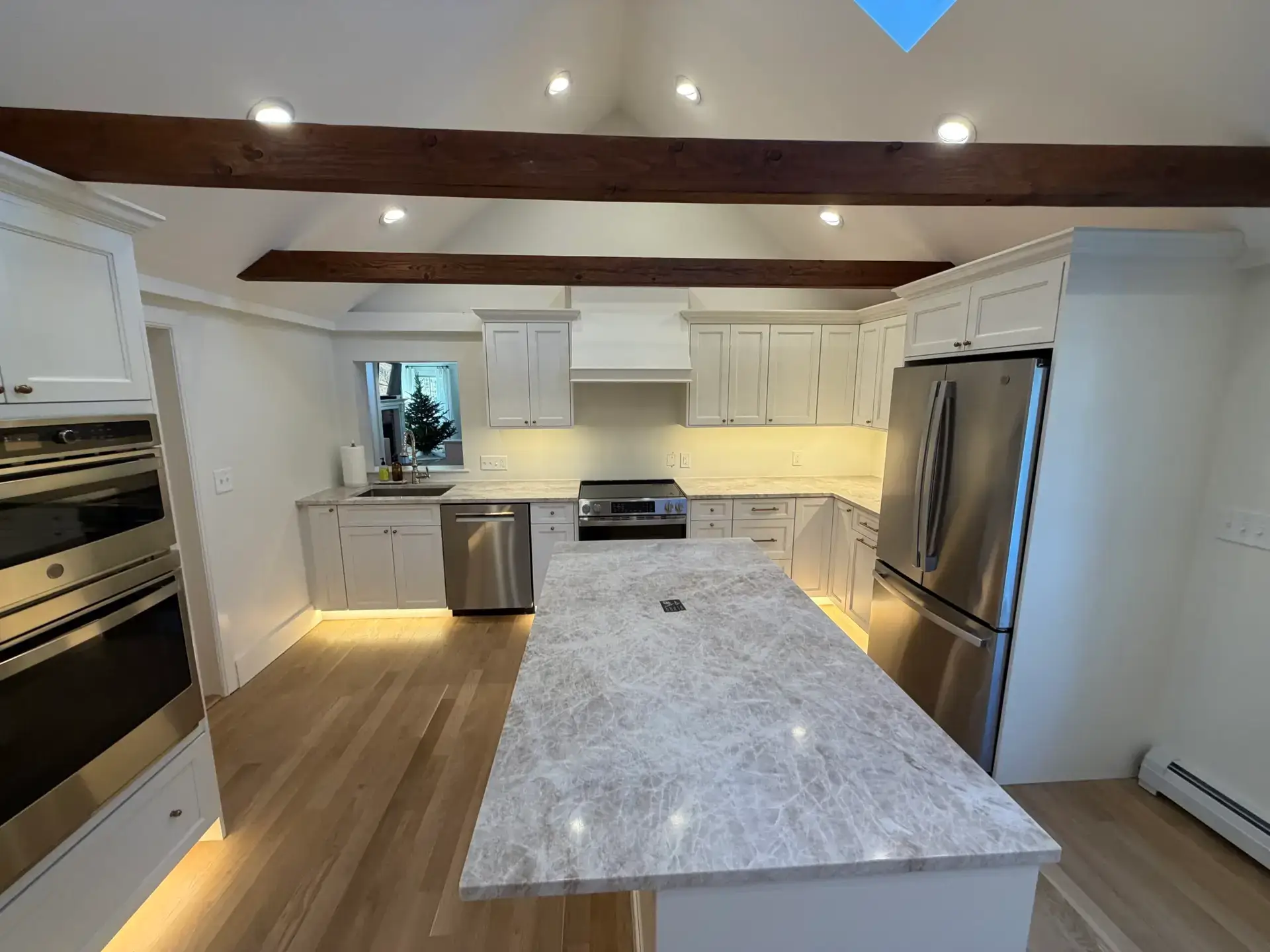 Modern kitchen design featuring white cabinetry, stainless steel appliances, and a large marble island, illuminated by recessed lighting and exposed wooden beams.