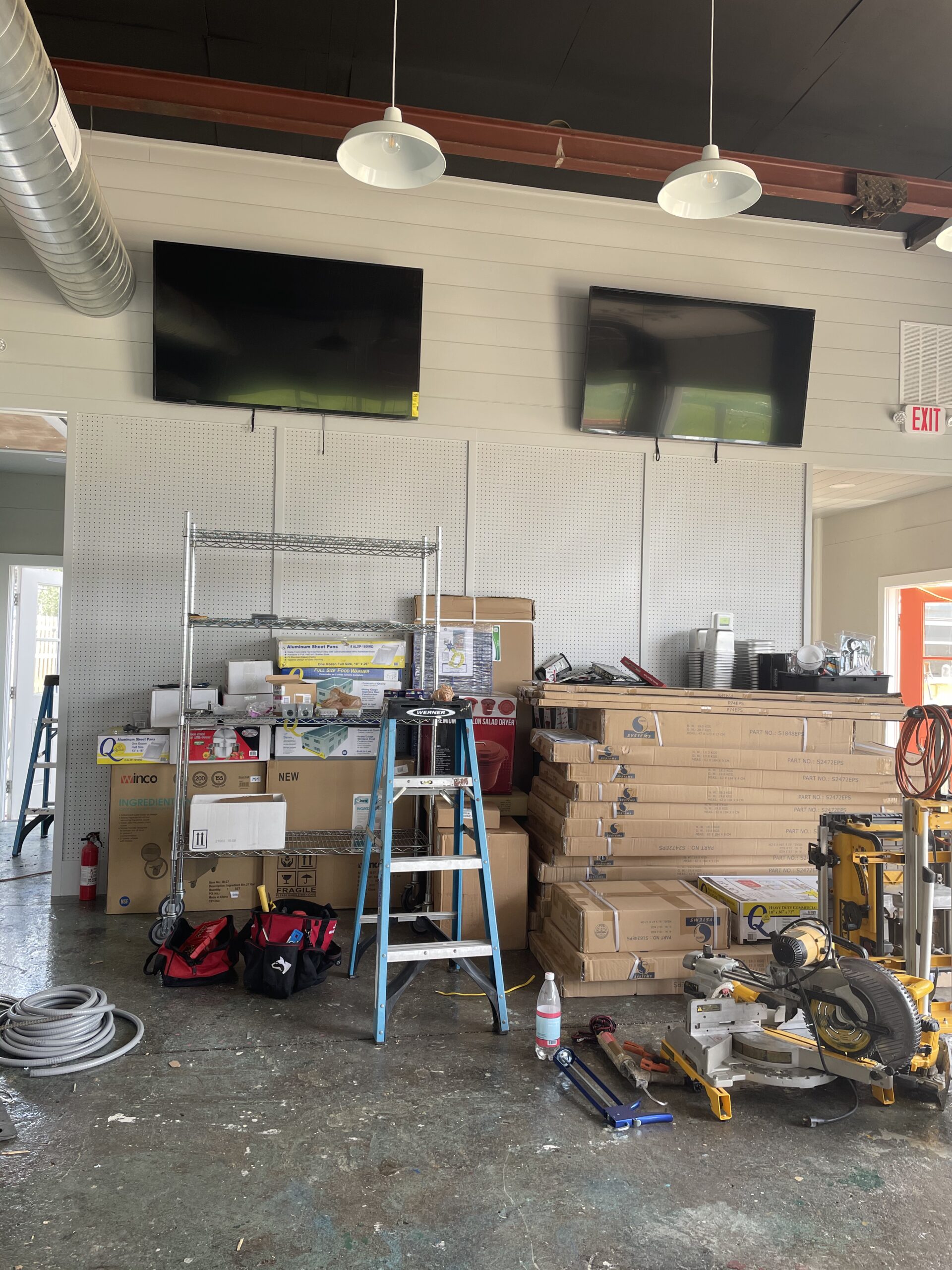 Interior workspace with two mounted TVs, shelving units, ladders, tools, and boxes, indicative of commercial electrical service preparations by Moore Electric Inc. in Plaistow, NH.