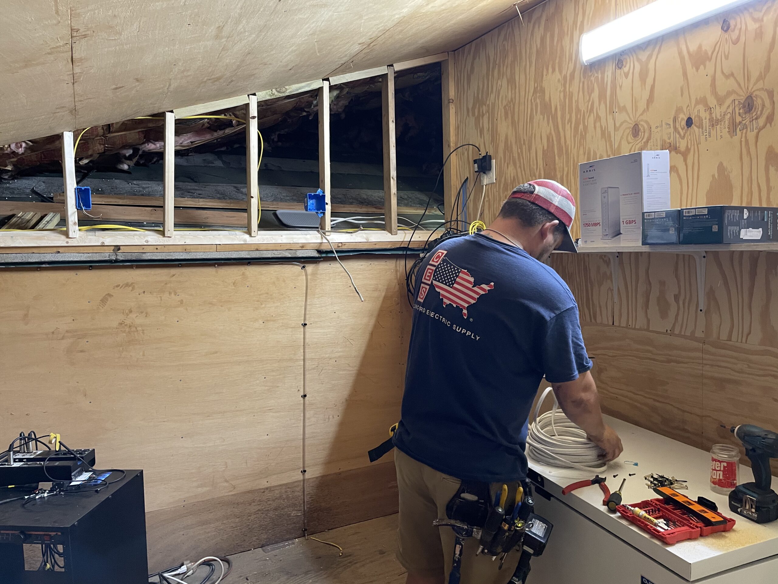 Electrician working on electrical wiring installation in a home, with tools and materials on a workbench, showcasing a partially constructed wall and overhead wiring.