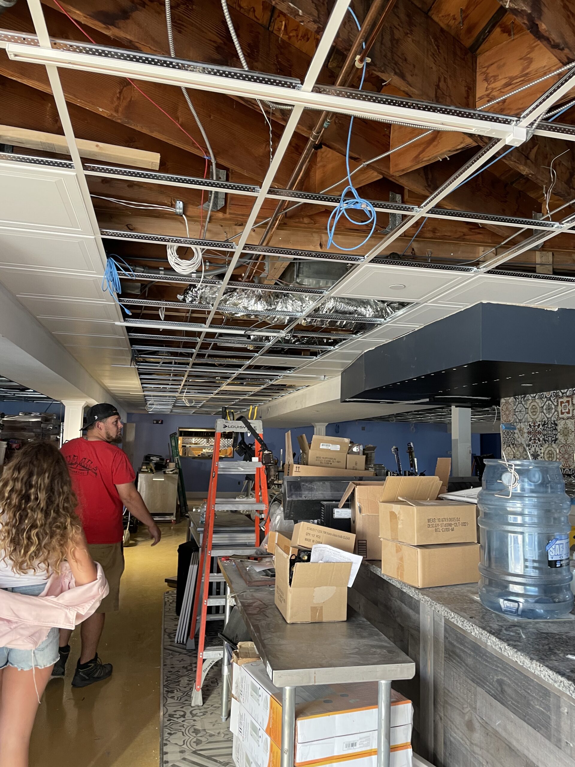 Interior view of a partially renovated space showing exposed wiring and ceiling framework, with a ladder and boxes, illustrating a kitchen renovation project related to electrical upgrades.