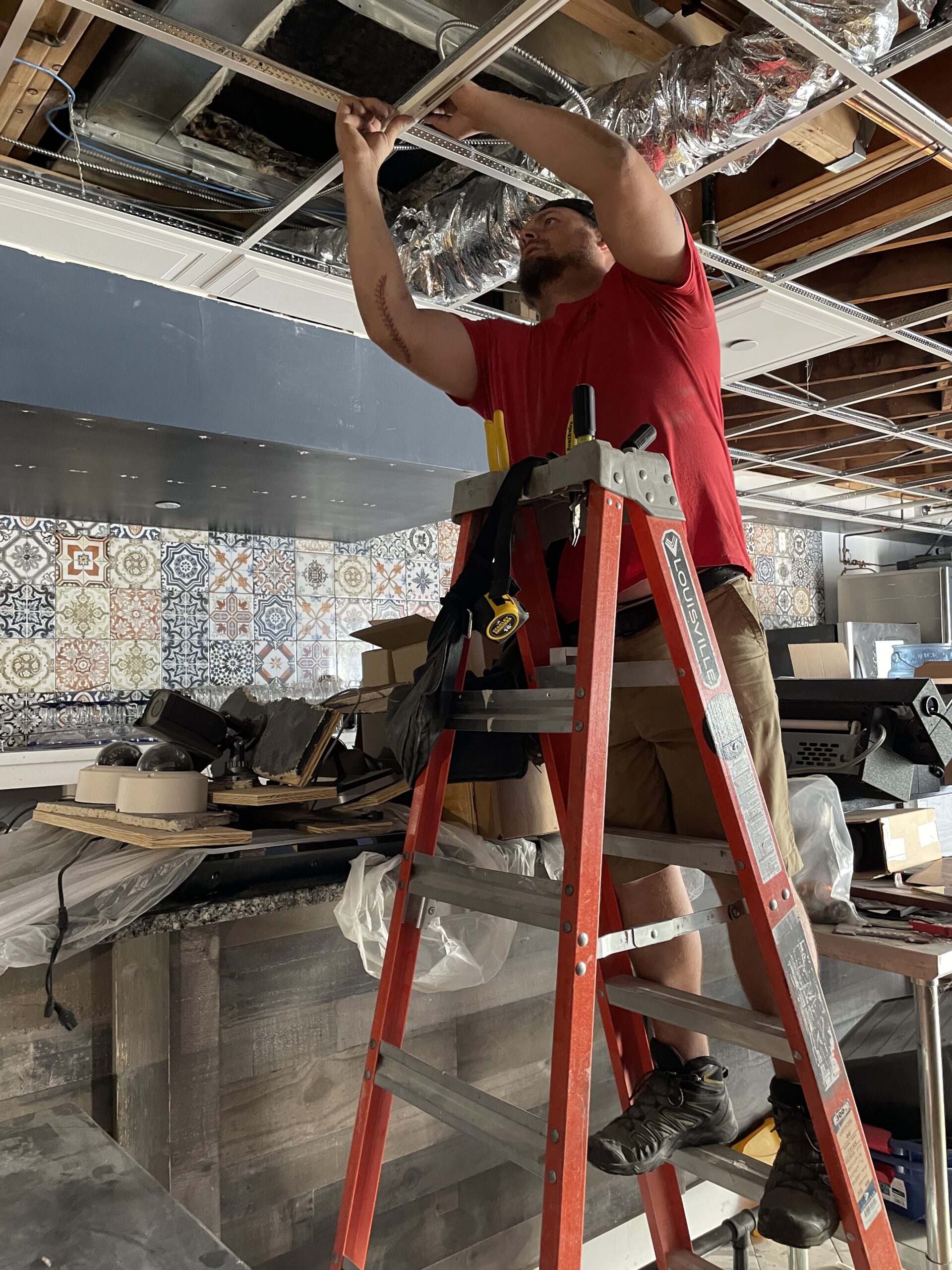 Electrician working on ceiling installation in commercial space, using a ladder, with tools and materials visible, showcasing electrical and lighting setup.