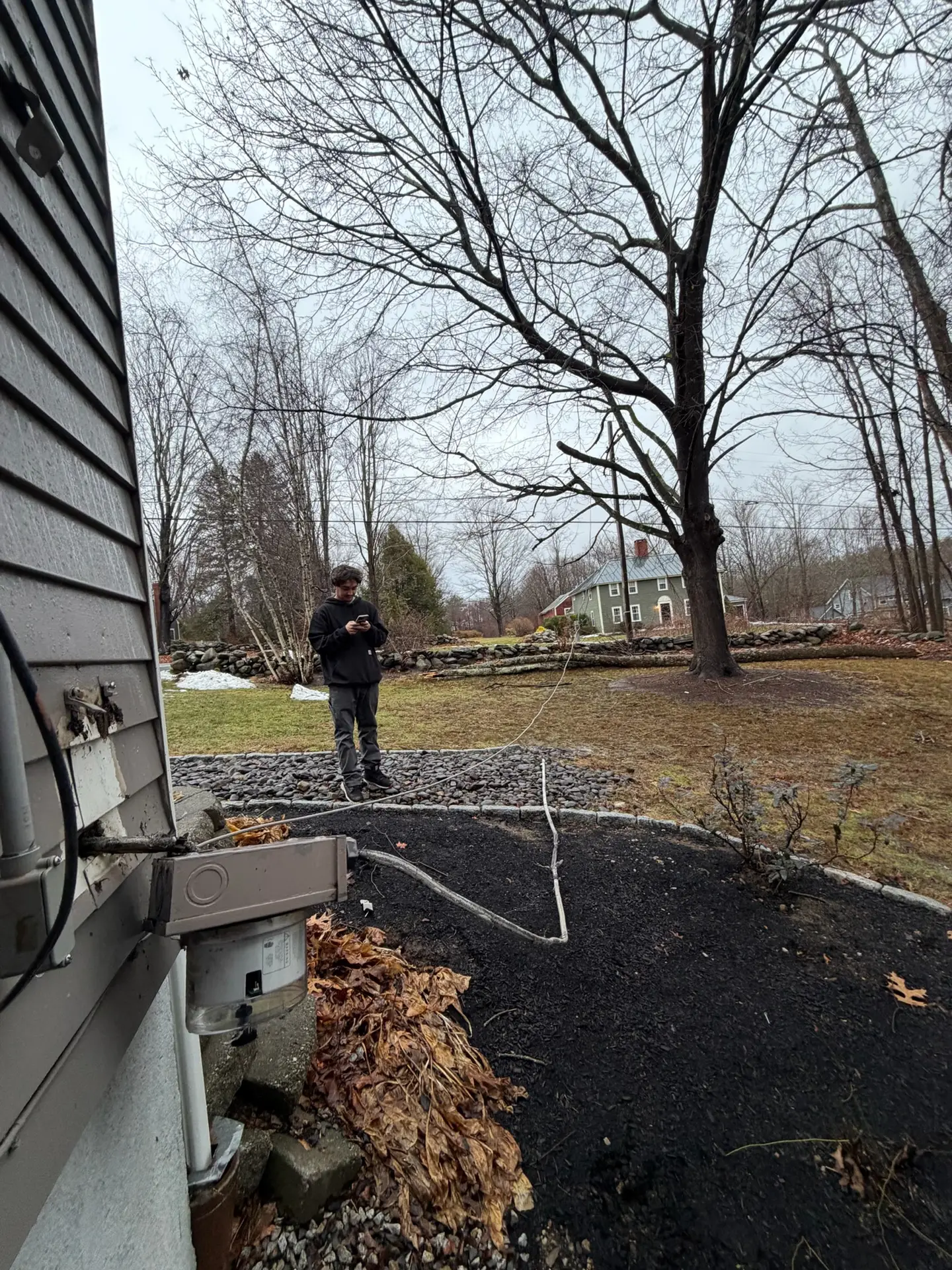 Man standing near a weatherhead and meter base, assessing potential electrical damage from storm-related issues, with fallen leaves and bare trees in the background.