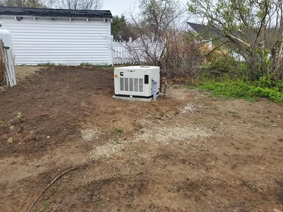 Generac standby generator installed in a cleared area, surrounded by sparse vegetation and a white building in the background, illustrating essential-circuit protection for homes in Plaistow, NH.
