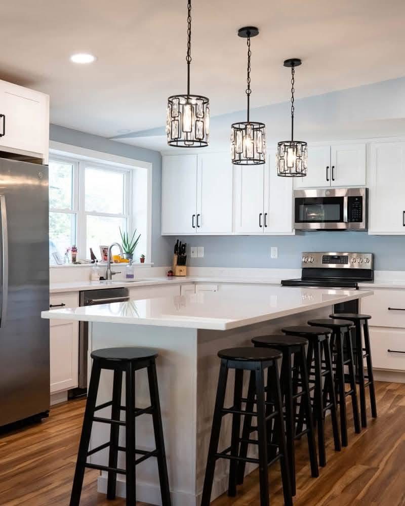 Modern kitchen with white cabinetry, stainless steel appliances, and stylish pendant lighting over a white countertop, featuring black bar stools, emphasizing renovation and electrical service expertise.