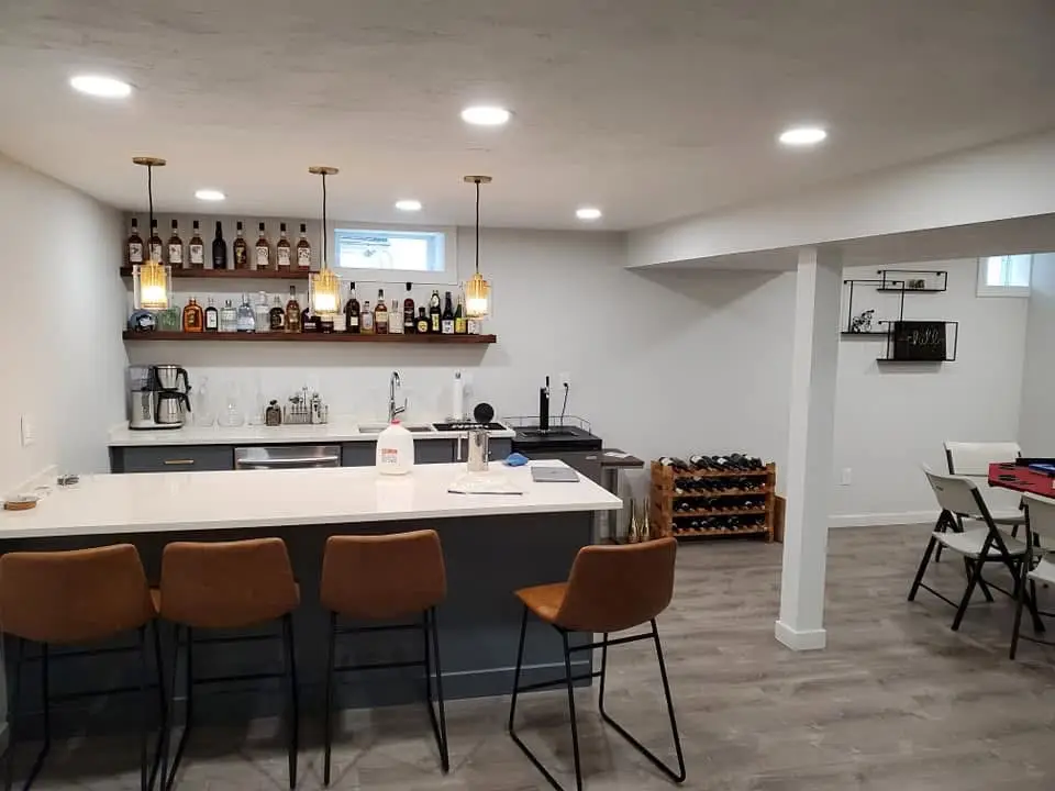 Modern basement bar area with illuminated pendant lights, stocked liquor shelf, sleek countertop, and seating, showcasing electrical work for kitchen and bath renovations by Moore Electric Inc.