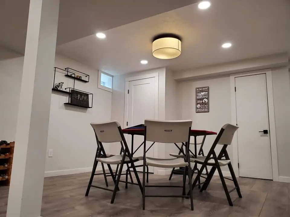 Basement dining area featuring a round table with four folding chairs, illuminated by a modern ceiling light fixture, showcasing newly installed lighting and a fresh, renovated space.