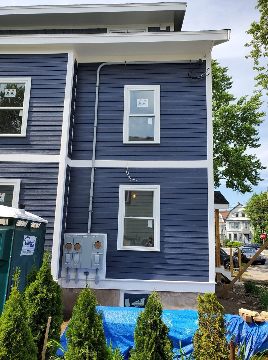 Newly installed electrical panel on blue residential siding, featuring three utility meters, surrounded by shrubs and construction materials, indicating ongoing electrical upgrades.
