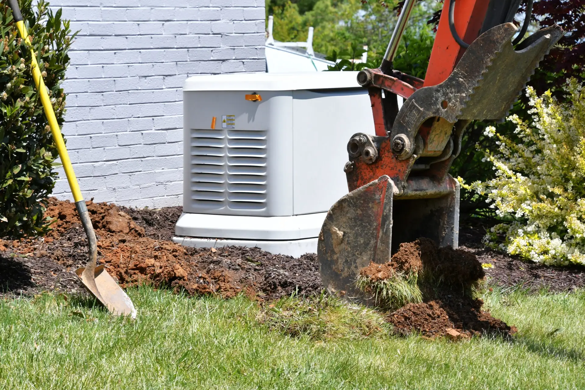 Backhoe digging dirt in lawn for generator installation, with a Generac unit visible, surrounded by greenery and mulch.