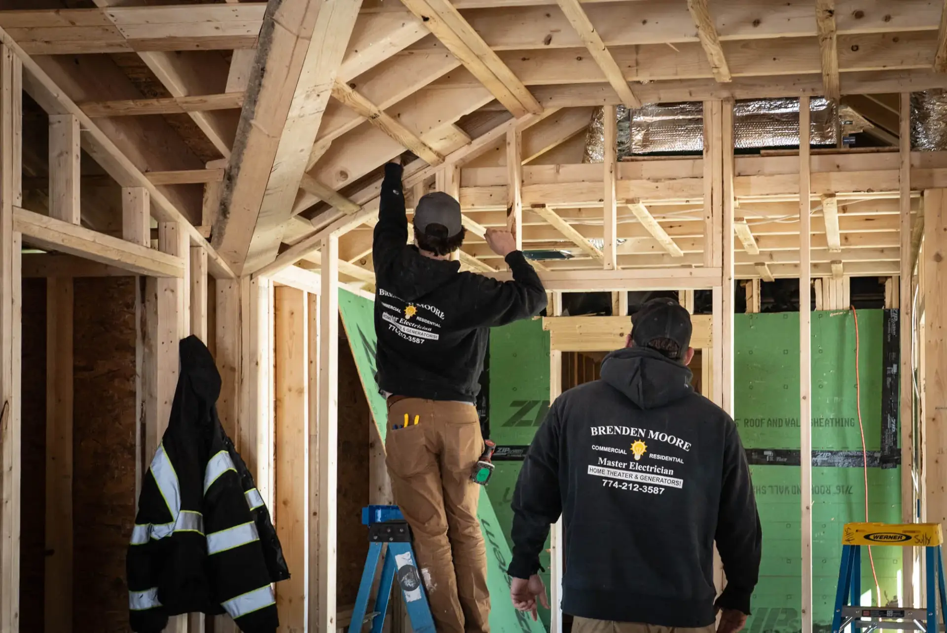 Electricians working on a construction site, installing electrical components, with visible branding on their hoodies, emphasizing expertise in electrical services.