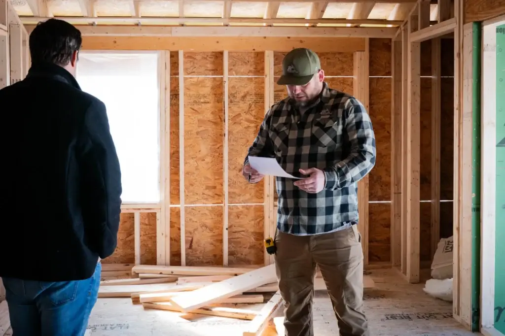 Two men discussing plans in a partially constructed room, one holding a document, showcasing a construction setting relevant to electrical service installations.