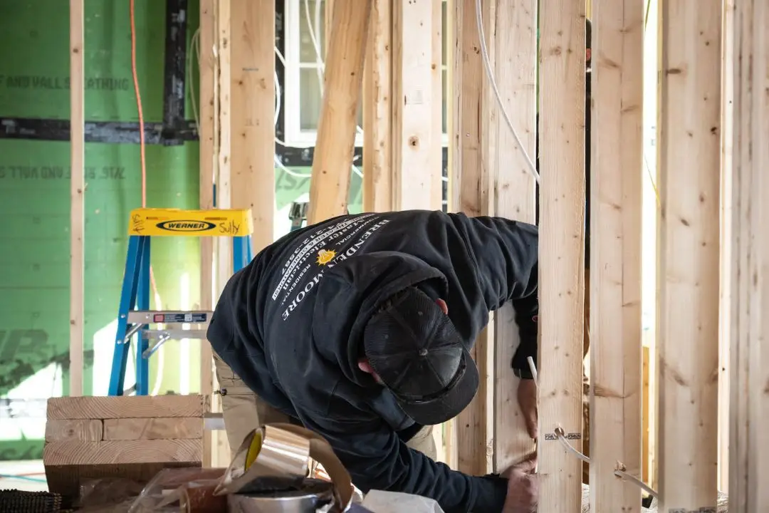 Electrician working on wiring installation in home addition, framed walls, tools nearby, ensuring compliance with electrical standards.